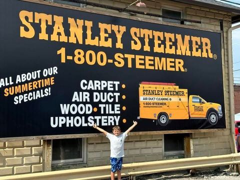 Child standing before a Stanley Steemer truck sign advertising carpet, air duct, wood, and tile upholstery cleaning. Child standing before a Stanley Steemer truck sign advertising carpet, air duct, wood, and tile upholstery cleaning.