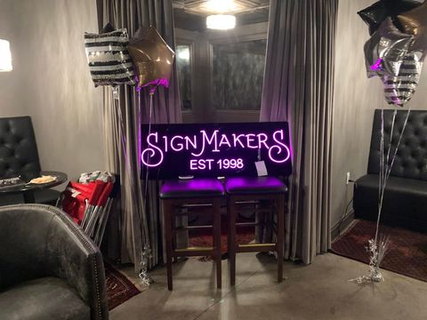 Neon pink “Signmakers EST 1996” sign in a room with two silver balloons and tables by curtains. Neon pink “Signmakers EST 1996” sign in a room with two silver balloons and tables by curtains.