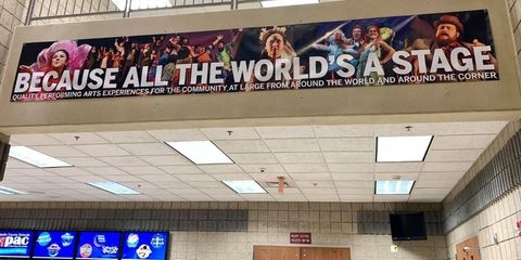 Banner in a hallway reads “Because all the world’s a stage” above ticket counters and ceiling lights. Banner in a hallway reads “Because all the world’s a stage” above ticket counters and ceiling lights.