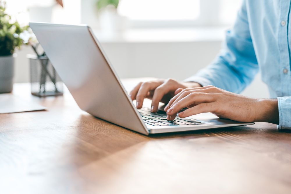 Person typing on a silver laptop on a wooden desk. Blue shirt, bright room, plant in background.