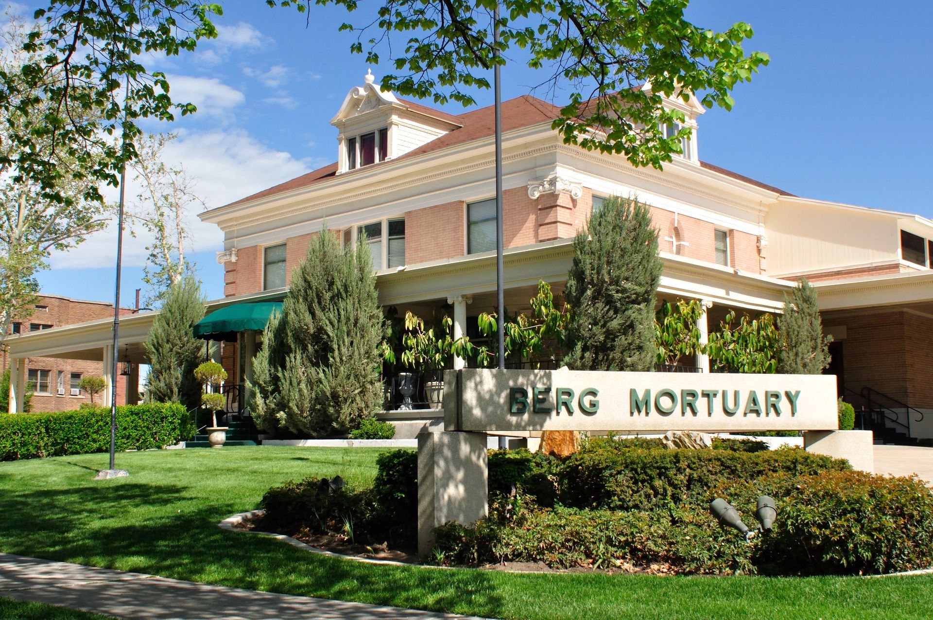 Berg Mortuary building with sign on a grassy lawn. Cream-colored exterior, brown roof, and blue sky.