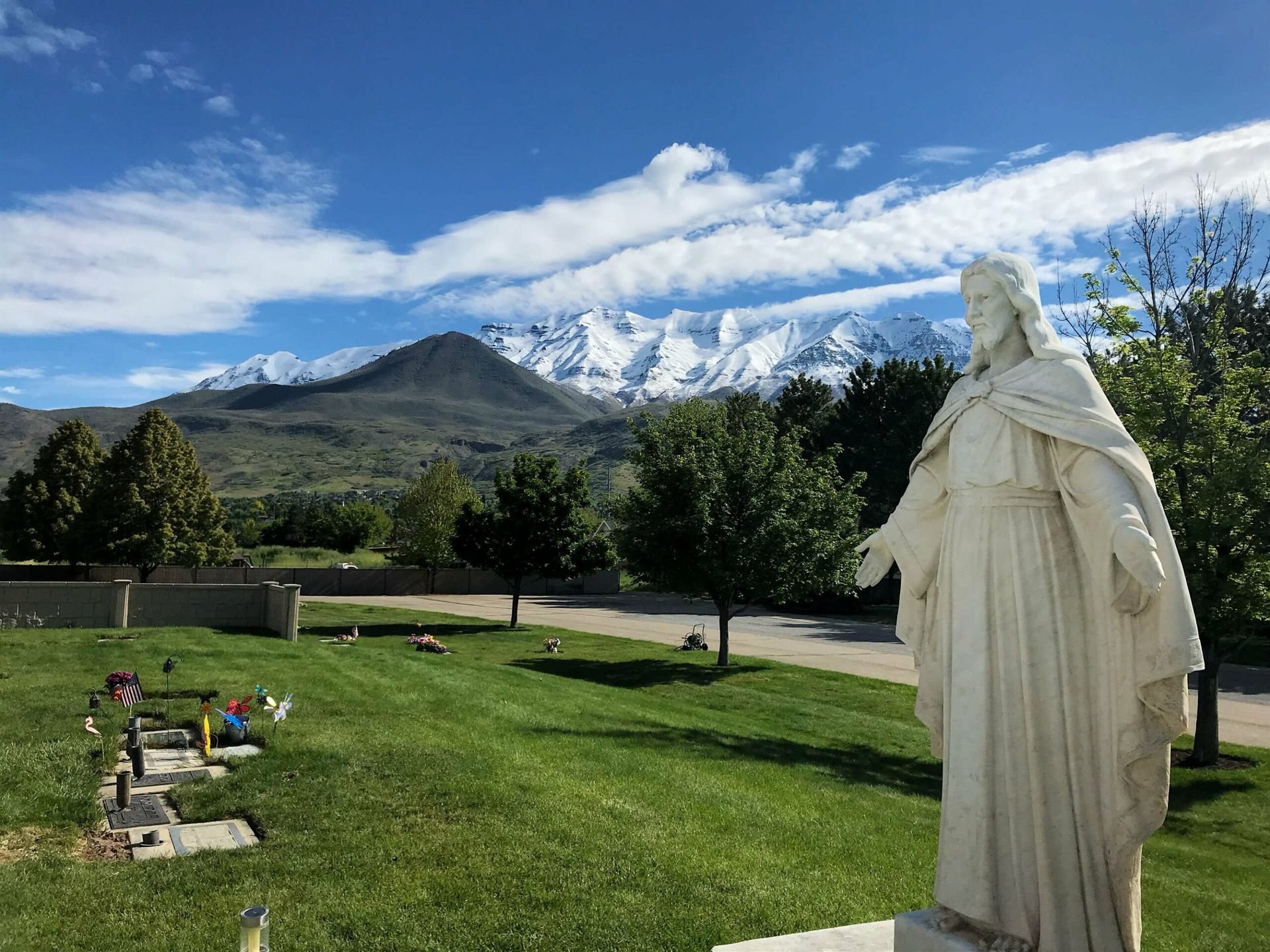 Statue of Jesus in a cemetery, snow-capped mountains in the background, green grass and blue sky.