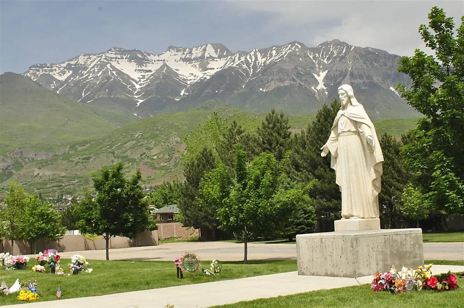 Statue of Jesus in a cemetery, snow-capped mountains in the background. Green trees and grass.