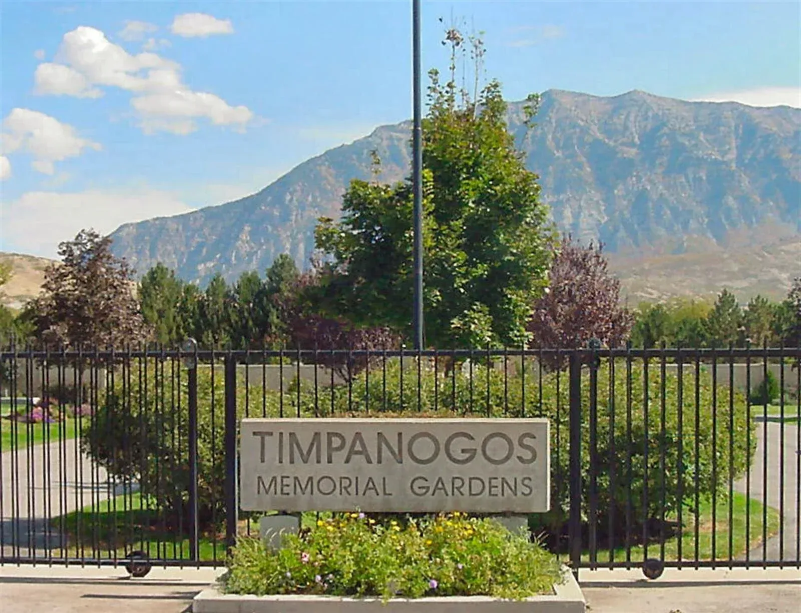 Sign for Timpanogos Memorial Gardens with mountains in the background. Black gate and green landscaping.