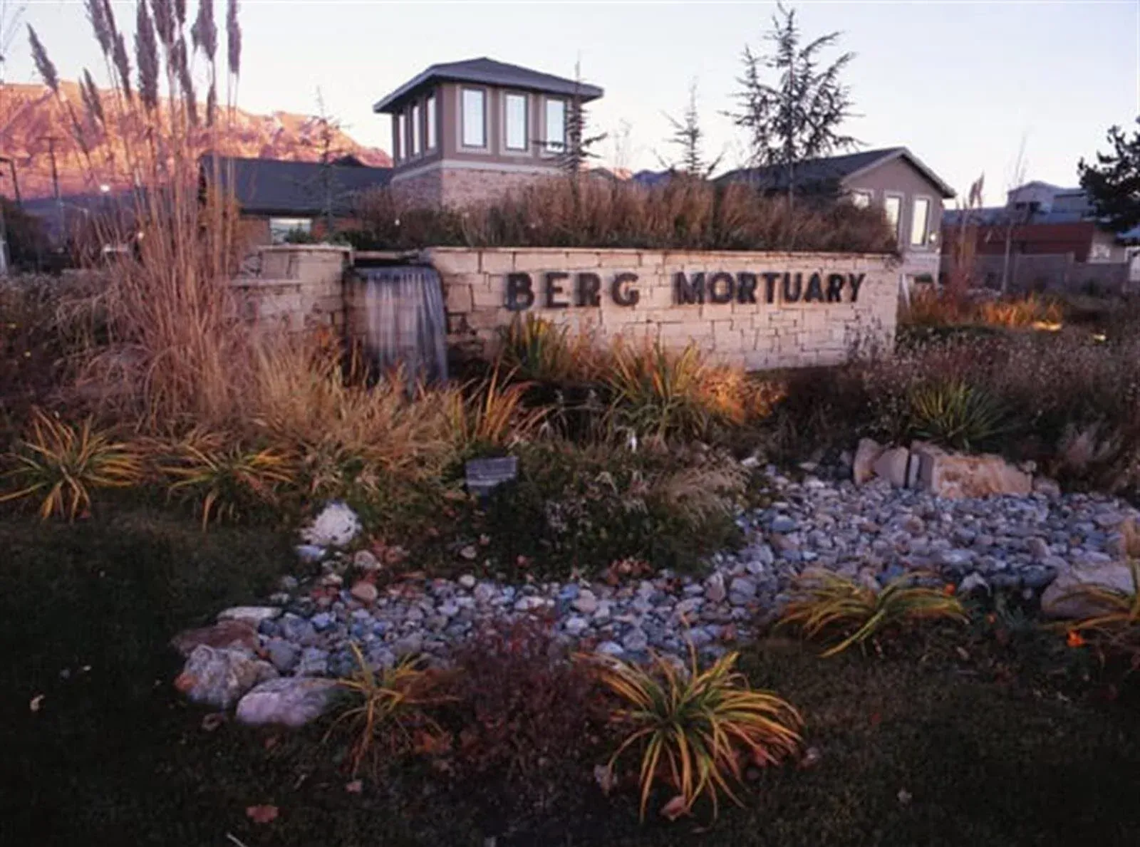 Berg Mortuary sign, surrounded by landscaping, small water feature, and building in background.