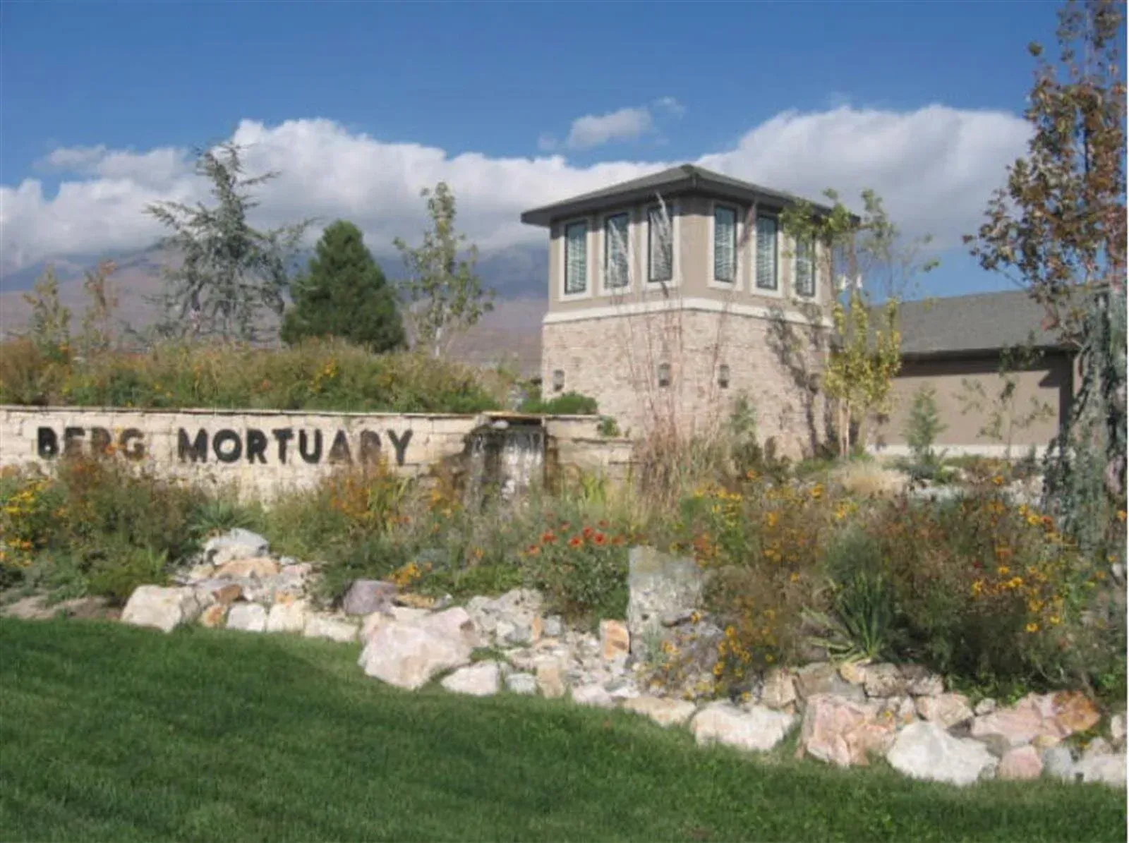 Berg Mortuary sign with stone building, flowers, and grass on a sunny day.