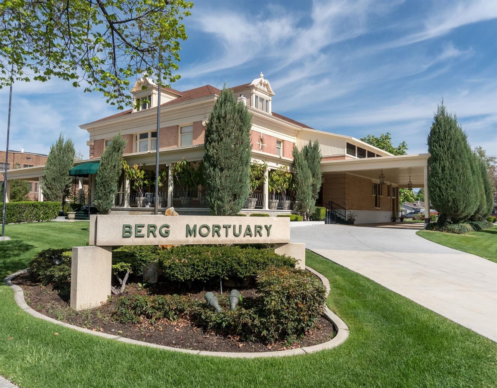 Berg Mortuary building with sign on a grassy lawn. Cream-colored exterior, brown roof, and blue sky.