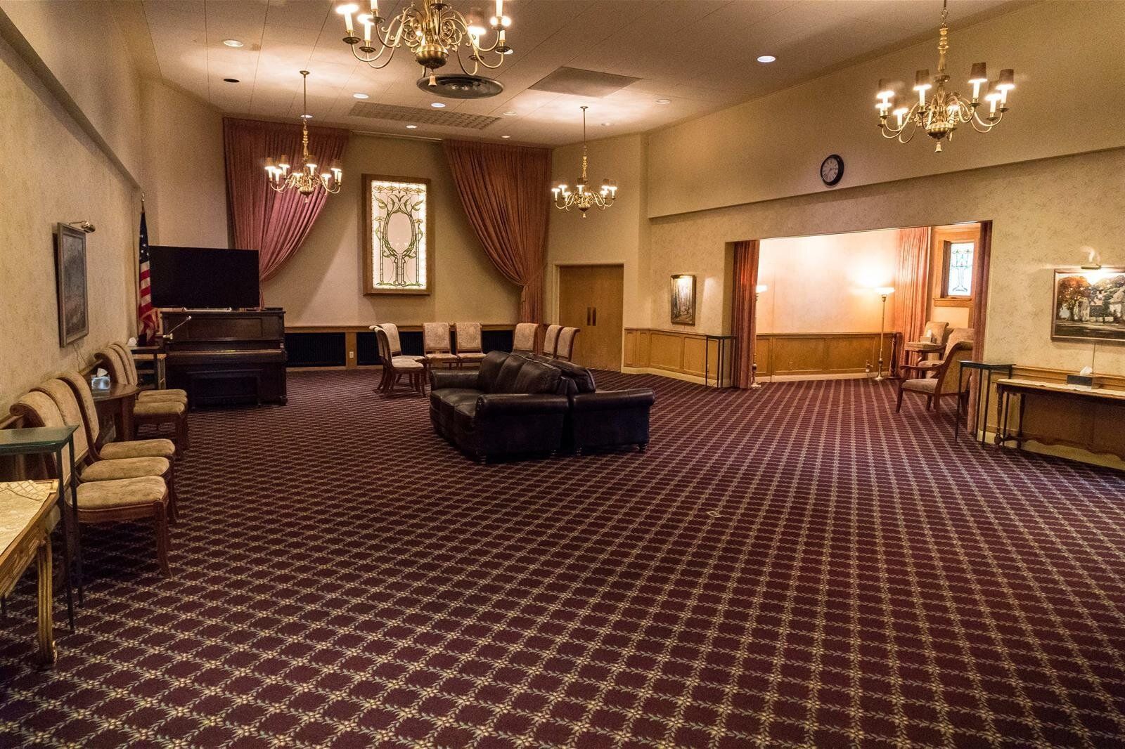 Large room, possibly a funeral home, with red patterned carpet, furniture, and chandeliers.