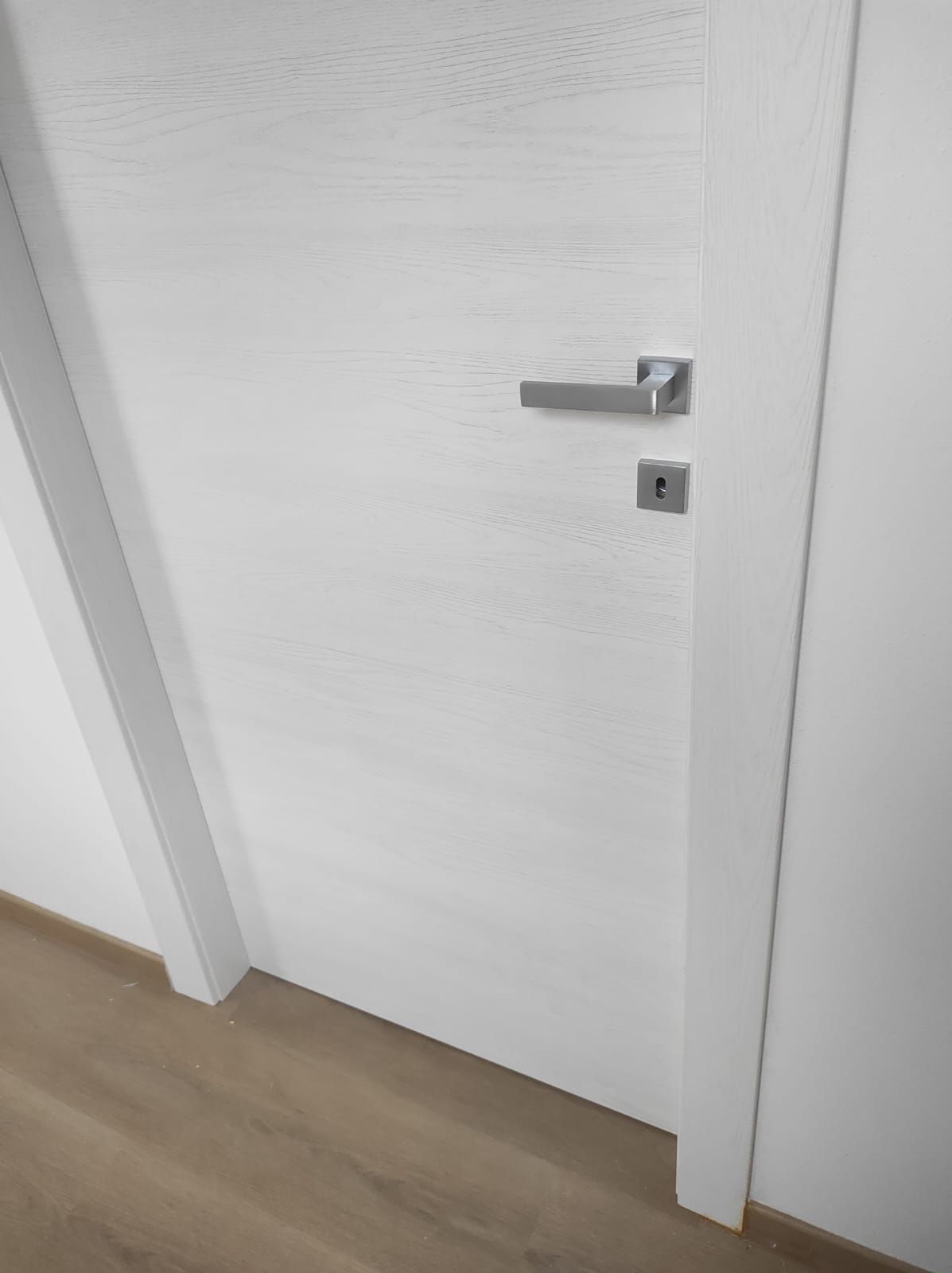 Una porta bianca con una maniglia argentata è appoggiata su un pavimento di legno.