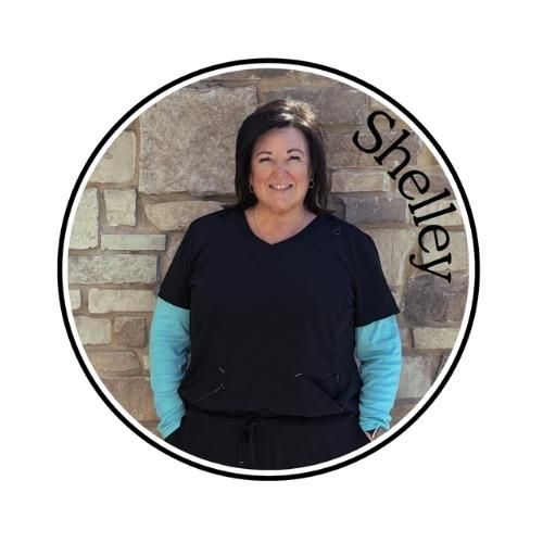 Woman standing against a stone wall, wearing a black top over turquoise sleeves, with