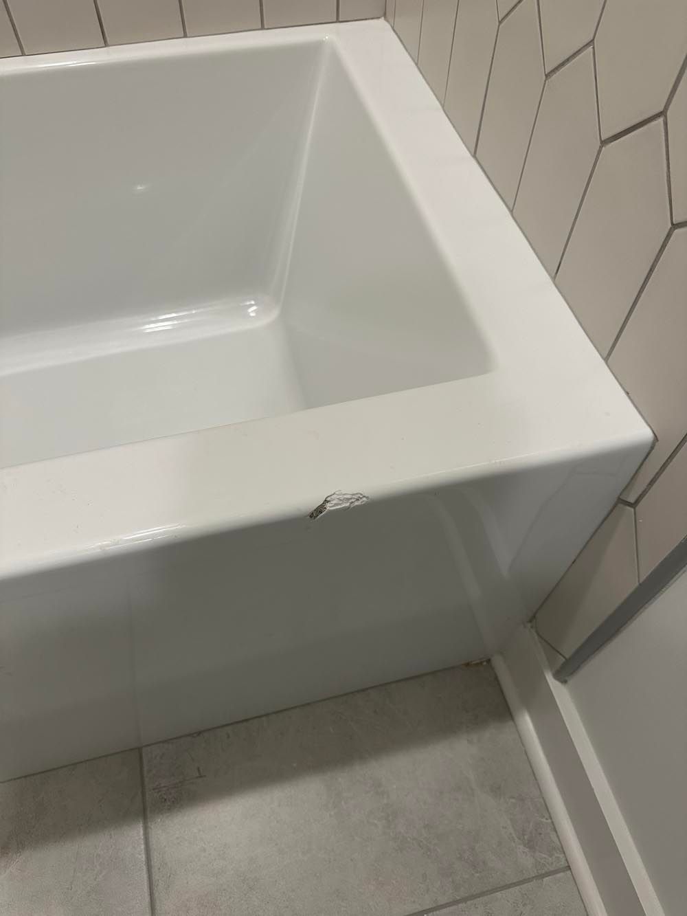 White bathtub with a chip on the front edge, set against light gray tiles and a tiled wall.