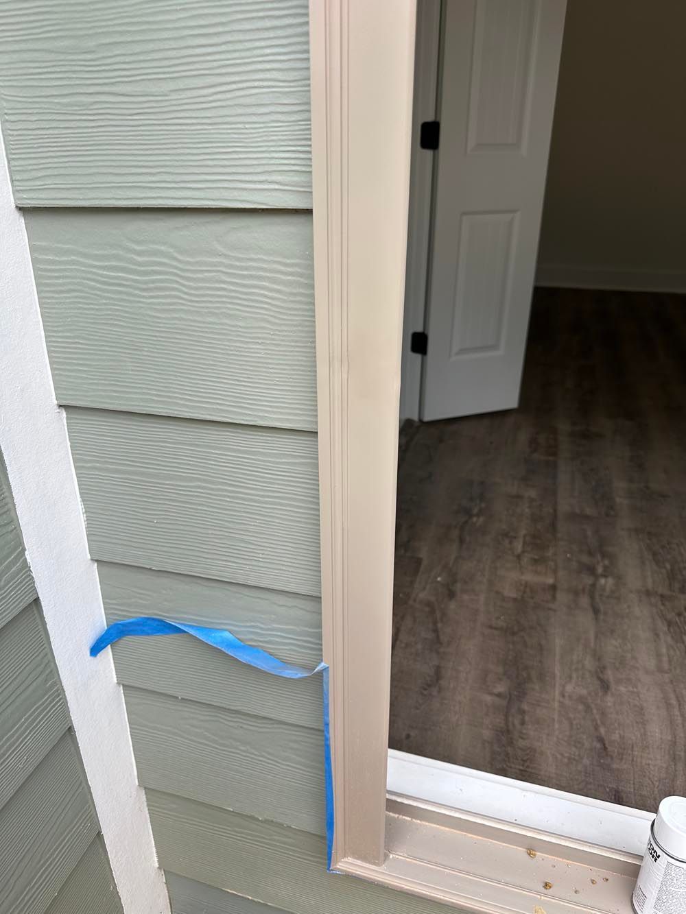 Exterior window frame with tan trim, blue tape, and green siding. Interior room with white door, and wood flooring.