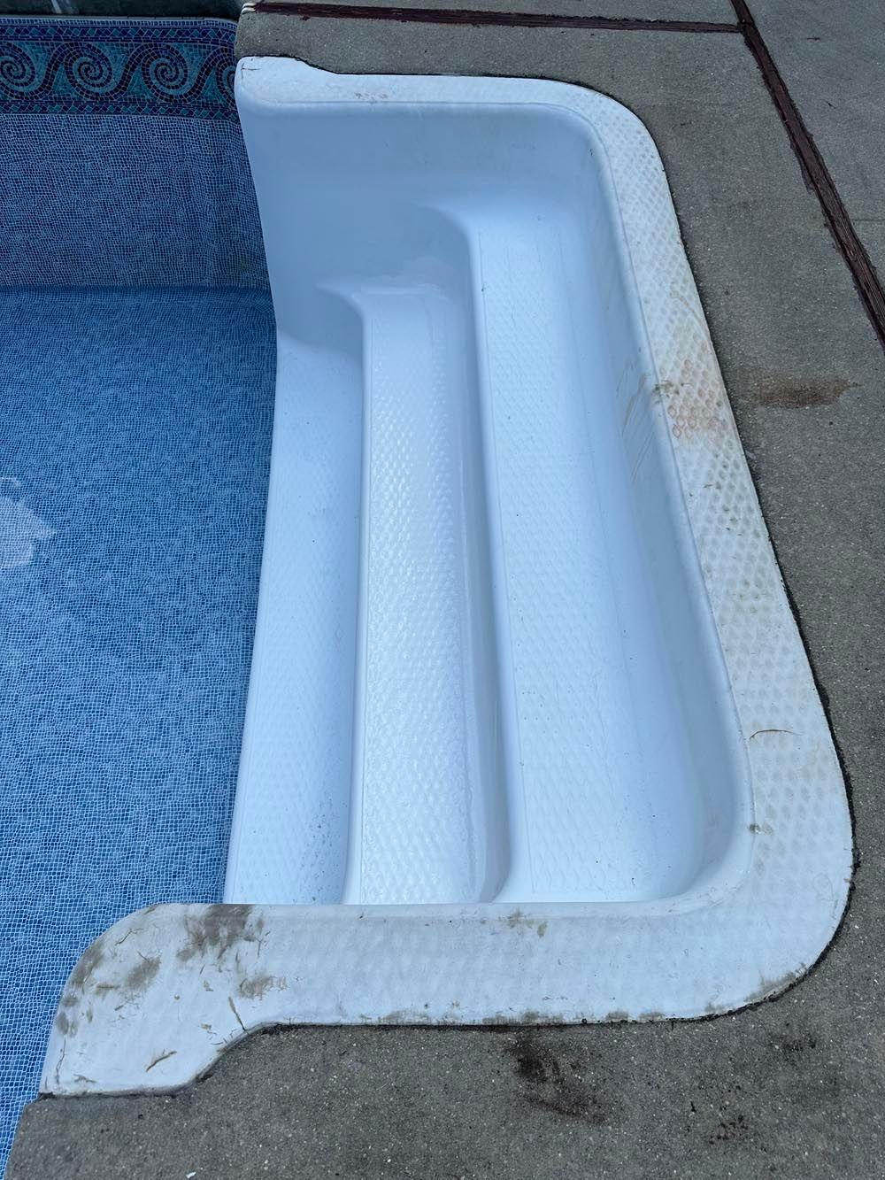 White in-pool steps, next to blue tile water, on a concrete pool deck.