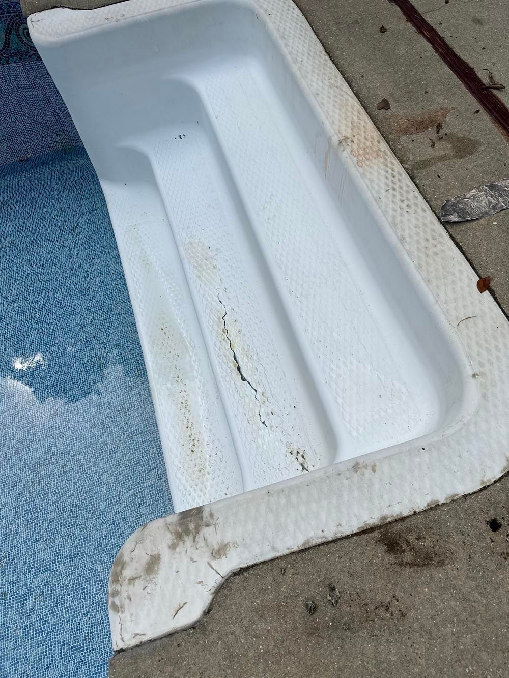 White pool steps with debris, set into a tiled pool with clear blue water. Concrete edge.
