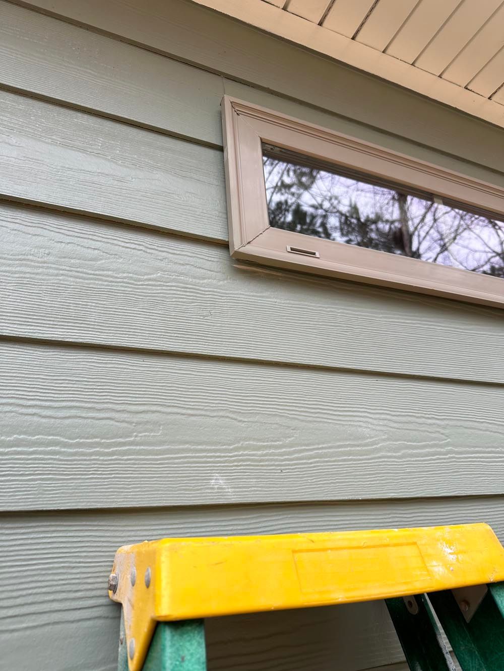 A ladder next to a green sided building with a brown-framed window.