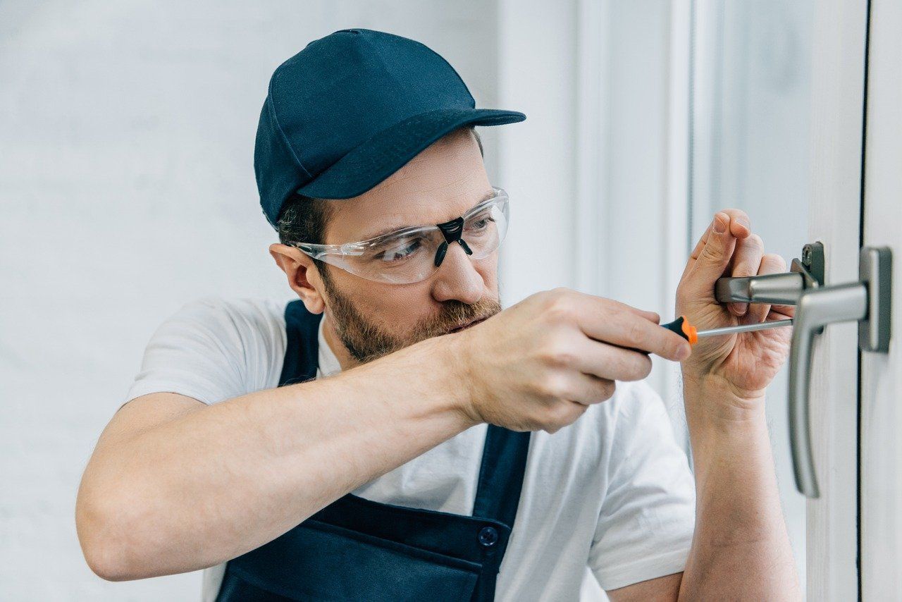 A man wearing safety glasses and a hat is fixing a door handle with a screwdriver.