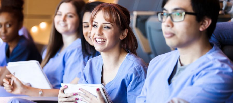 Medical students in scrubs, using tablets, smiling in a classroom.