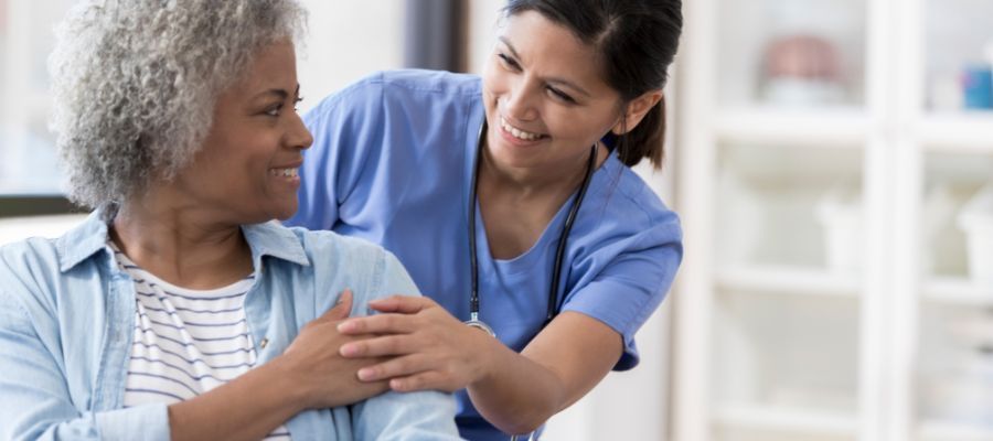 A healthcare worker in blue scrubs puts a supportive hand on an elderly woman's arm, both smiling.