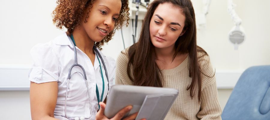 A doctor and patient look at a tablet in a medical setting. The doctor wears a stethoscope and smiles.