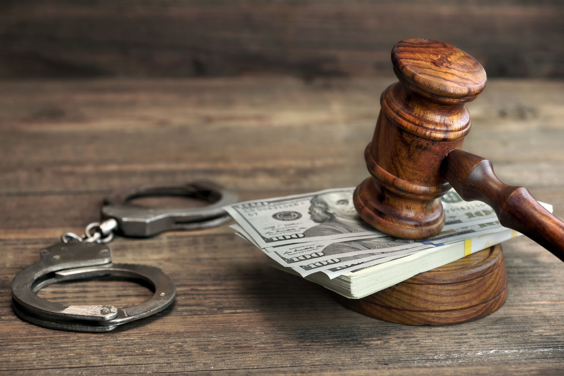 A wooden judge 's gavel with a stack of money and handcuffs on a wooden table.