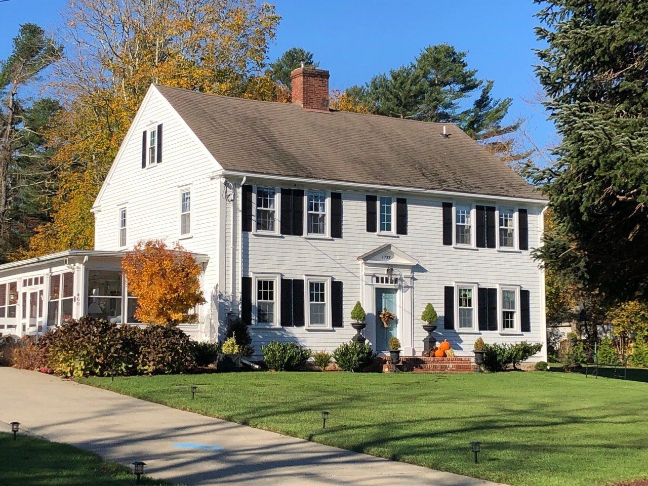 White two-story house with black shutters, brick chimney, and a sunroom, on a green lawn with trees.
