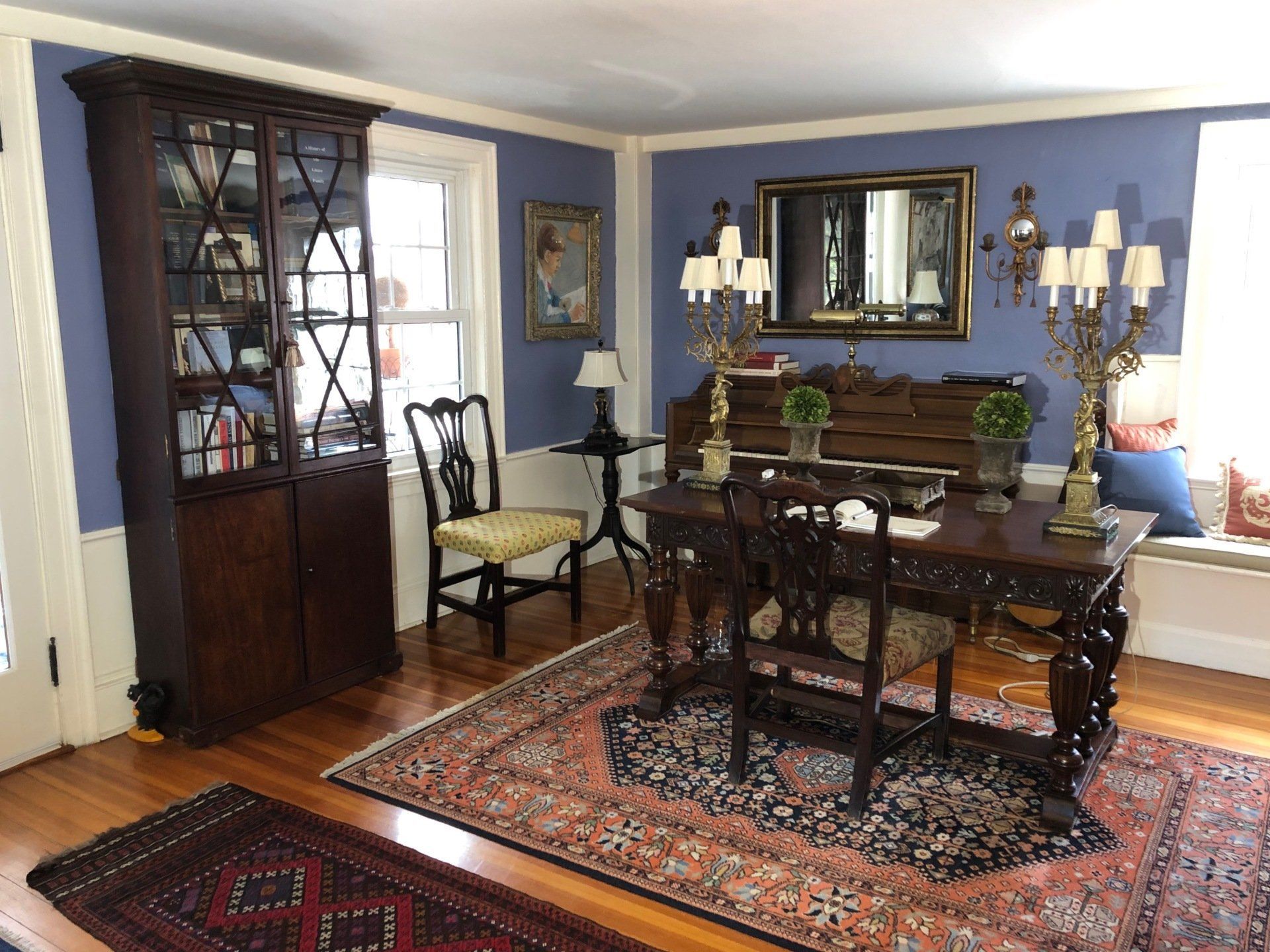 Antique study with blue walls, ornate desk and chairs, Persian rugs, and a tall wooden bookcase.