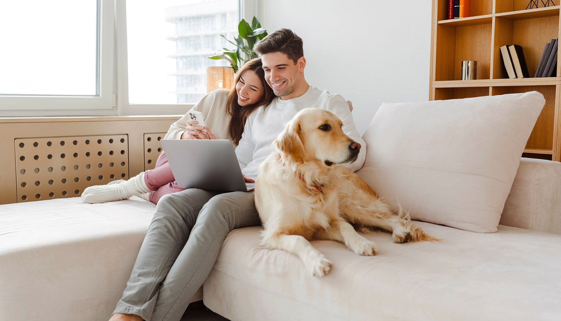 A man and woman are sitting on a couch with a dog and a laptop.