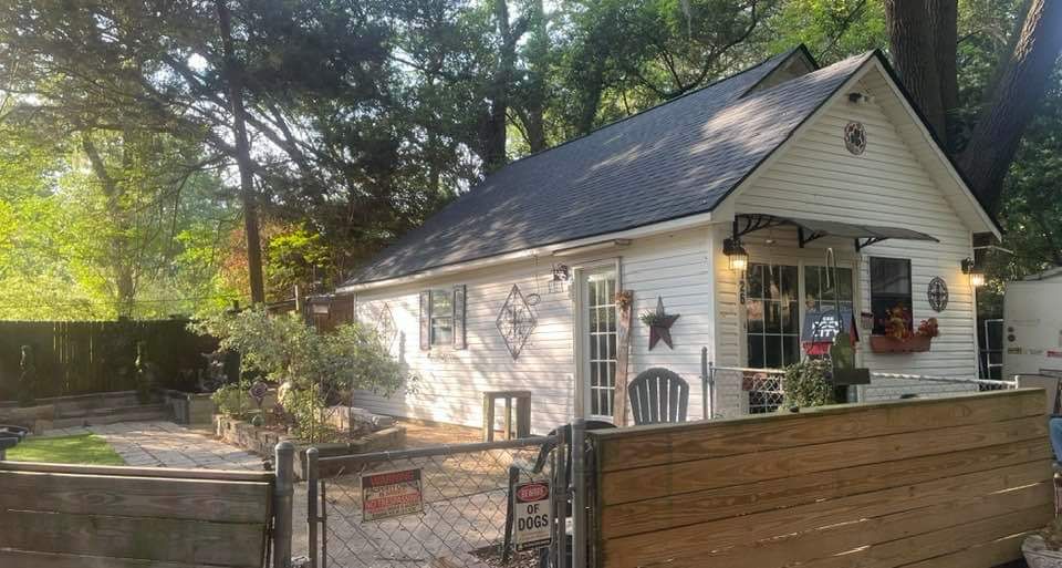 A small white house with a black roof is surrounded by trees and a wooden fence.