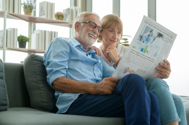 An elderly couple is sitting on a couch reading a newspaper.