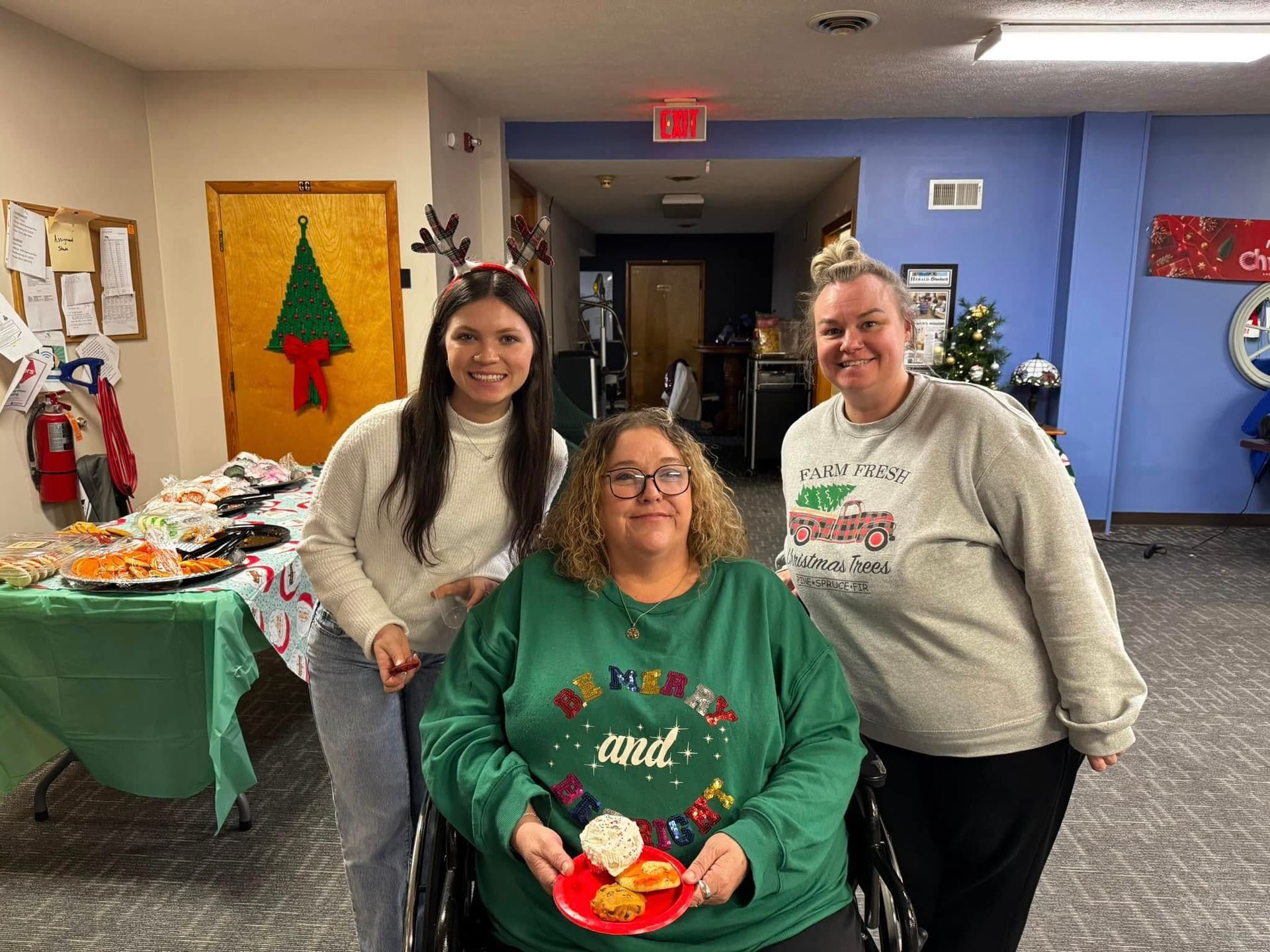 Three women are posing for a picture in a room with a woman in a wheelchair holding a plate of food.
