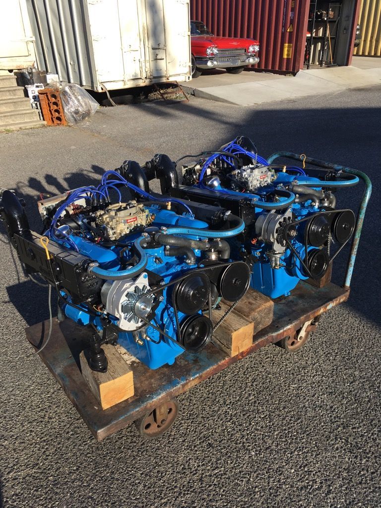 Two blue marine engines on a metal cart, outdoors with shipping containers and a classic red car in the background.