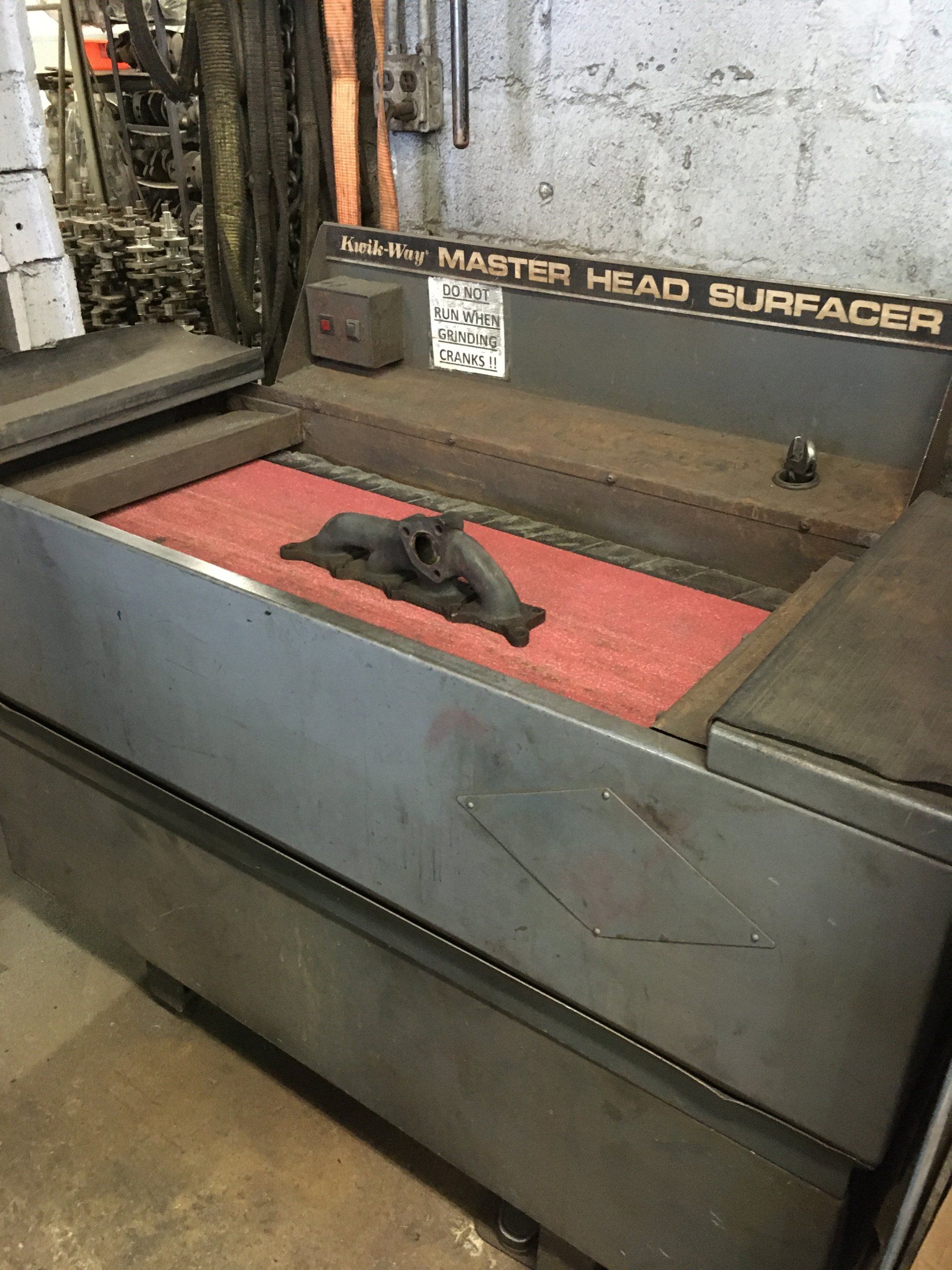 A gray Head Surfacer machine with a black car part on a red pad inside a shop.