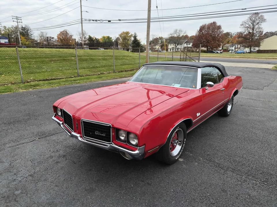 Red 1970 Oldsmobile Cutlass convertible on asphalt. Black top is up; the car is parked in front of a grassy hill.
