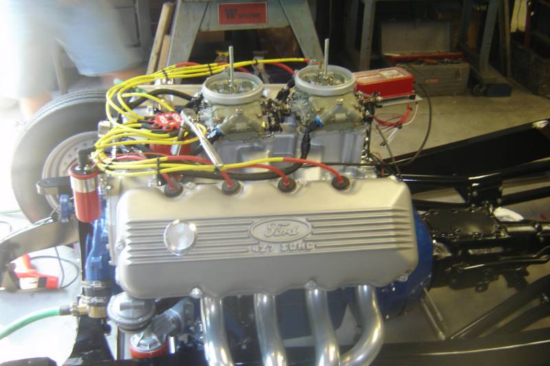 Ford engine with dual carburetors, silver valve covers, blue block, yellow wires, on a classic car chassis.