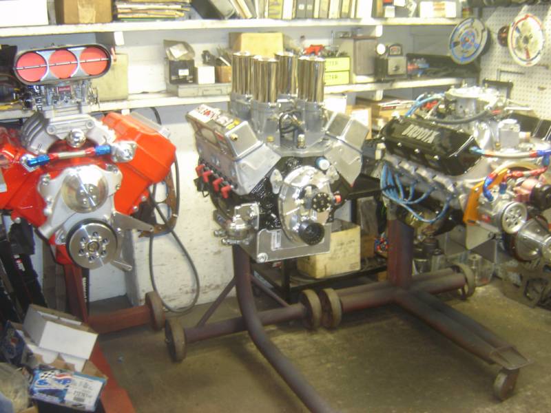 Three high-performance car engines on stands, brightly colored with various chrome components, in a cluttered workshop.