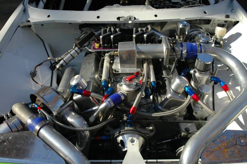 Engine bay of a white race car with numerous chrome tubes, blue accents, and silver components.