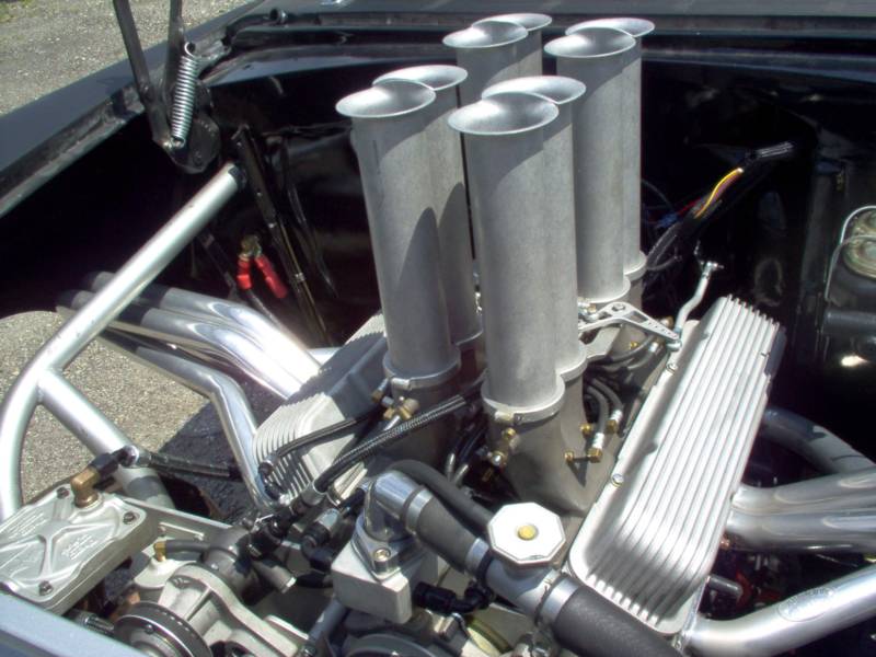 Close-up of a car engine with six tall intake tubes, gray and chrome components, and a roll cage.