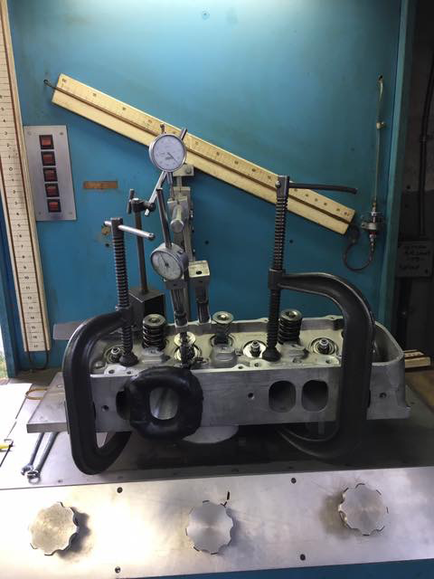 Cylinder head clamped in a machine with gauges, likely for valve seat work. Light blue wall.