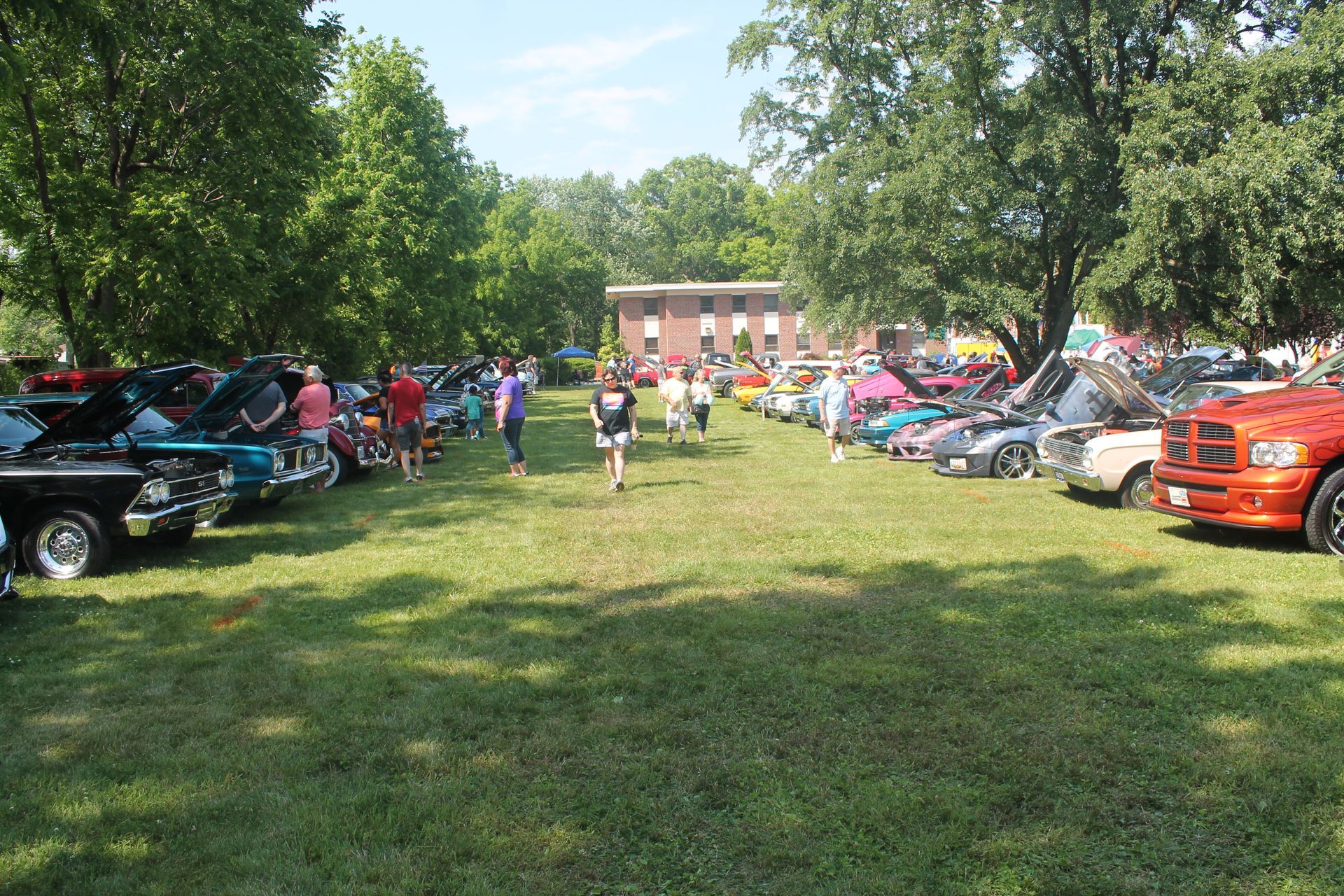 Classic cars on display at outdoor event; people walking between rows. Sunny day.