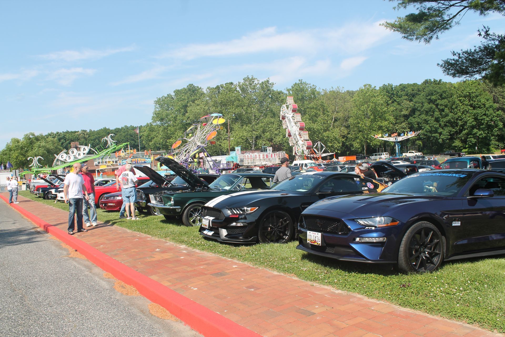 Cars displayed at a car show on a sunny day, people looking at the cars; amusement park rides in background.
