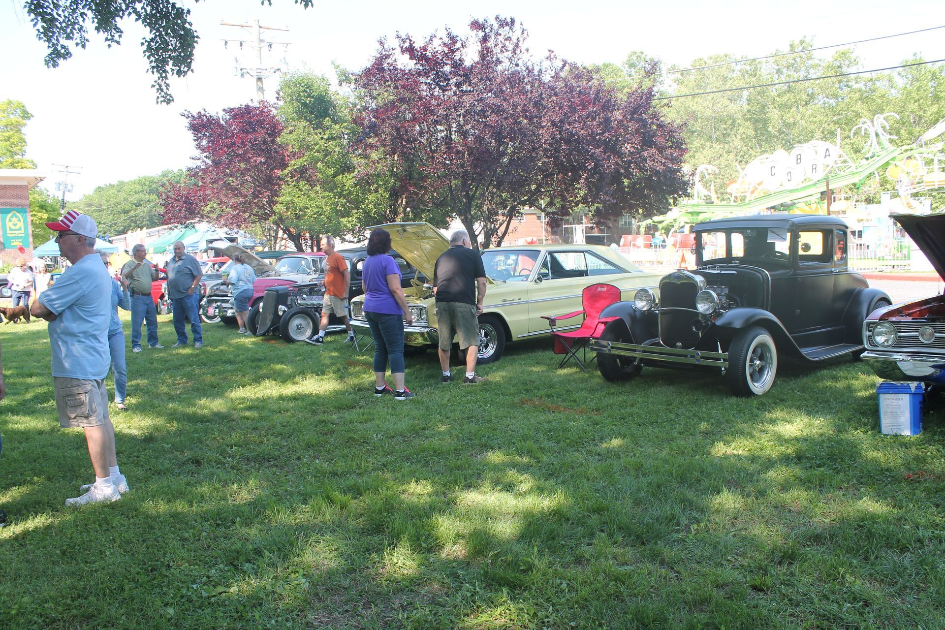Classic cars on display at an outdoor event with people viewing them. Green grass, trees, and sunny weather.