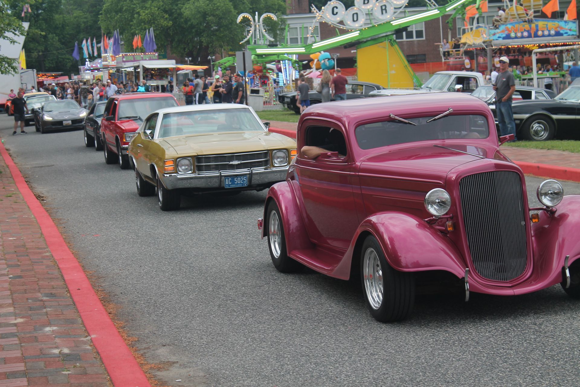 Classic cars parade down a street lined with people and carnival rides.