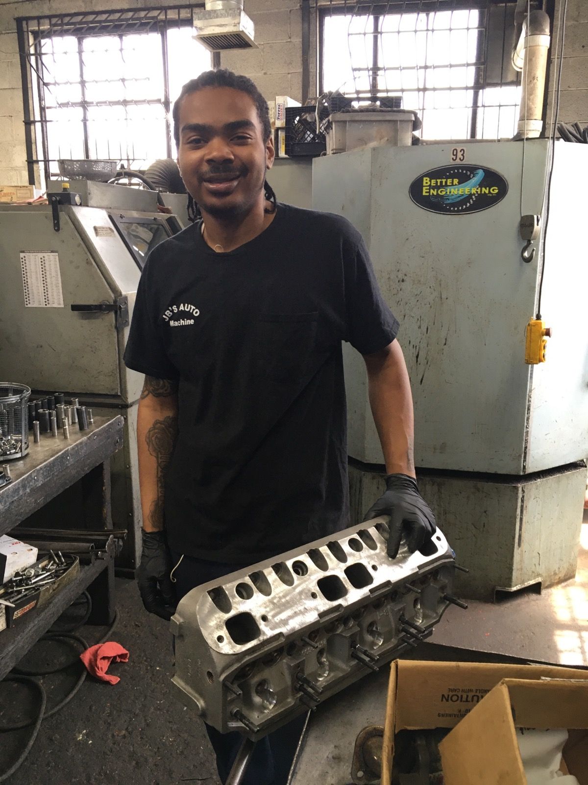 Smiling worker in a workshop holding a metal engine part beside machinery and boxes.