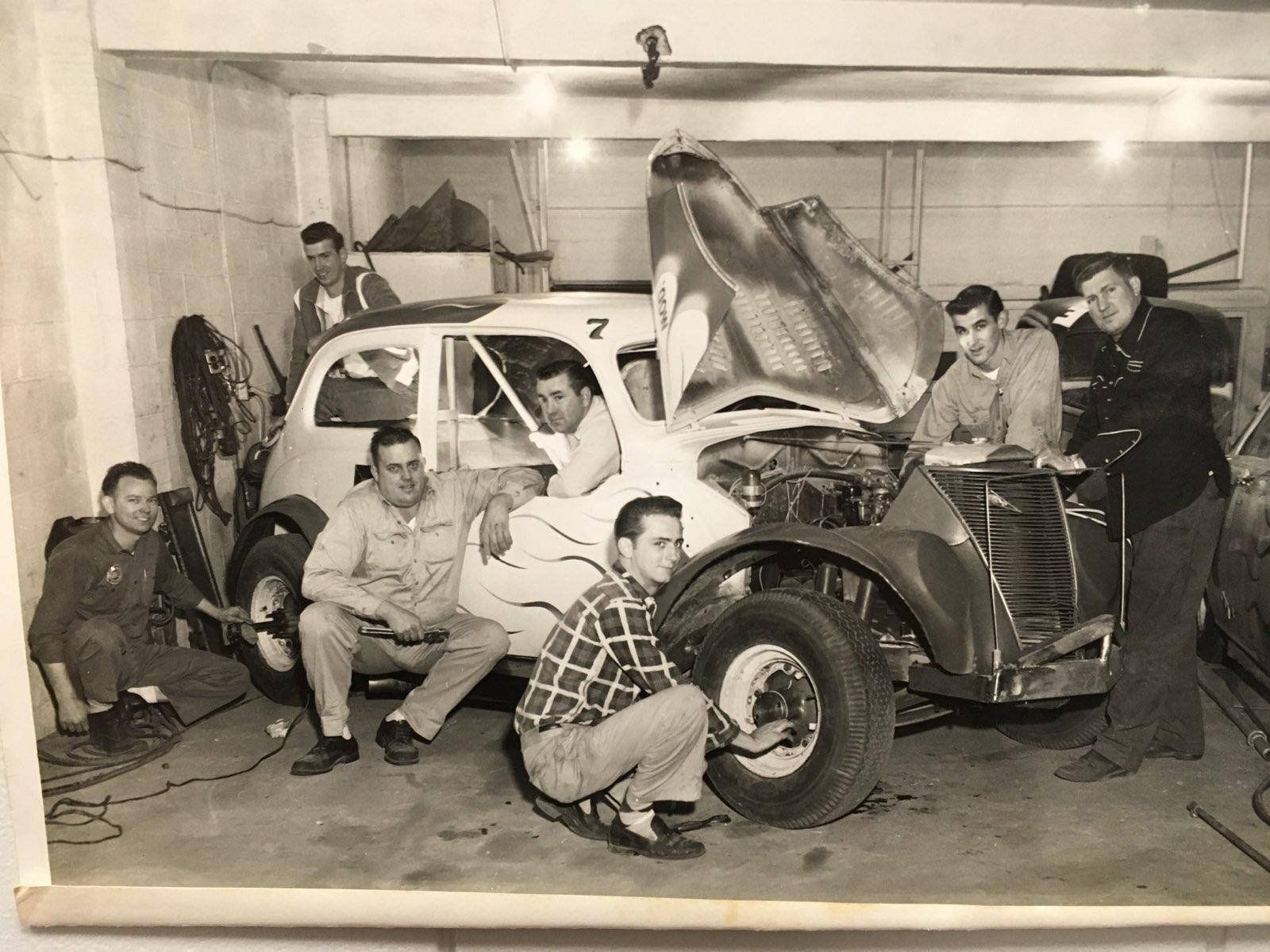 Men surrounding and working on a vintage race car in a garage. The car's hood is up, and they are making repairs.