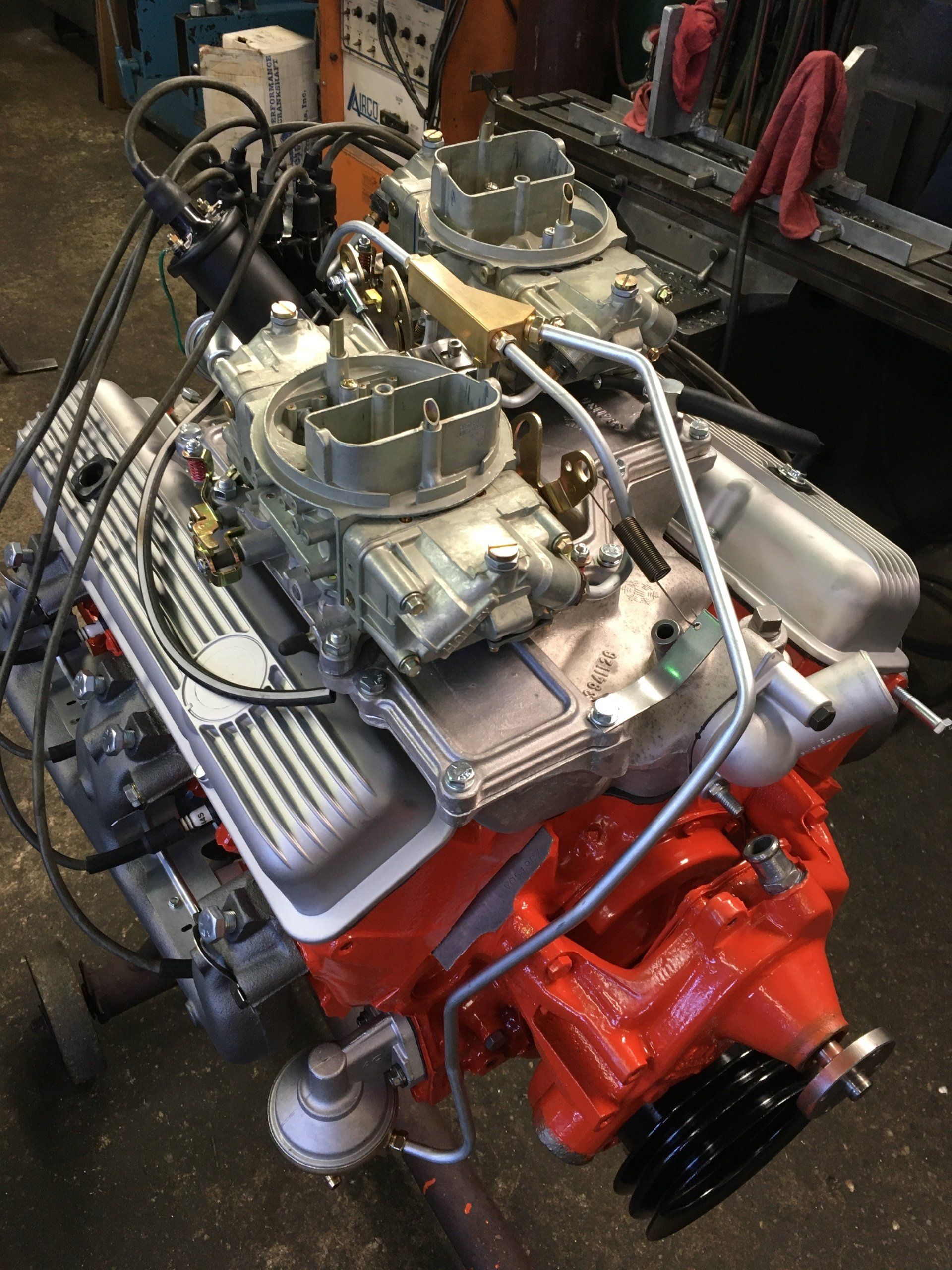 A vintage orange engine with silver carburetors, and valve covers sits in a workshop.