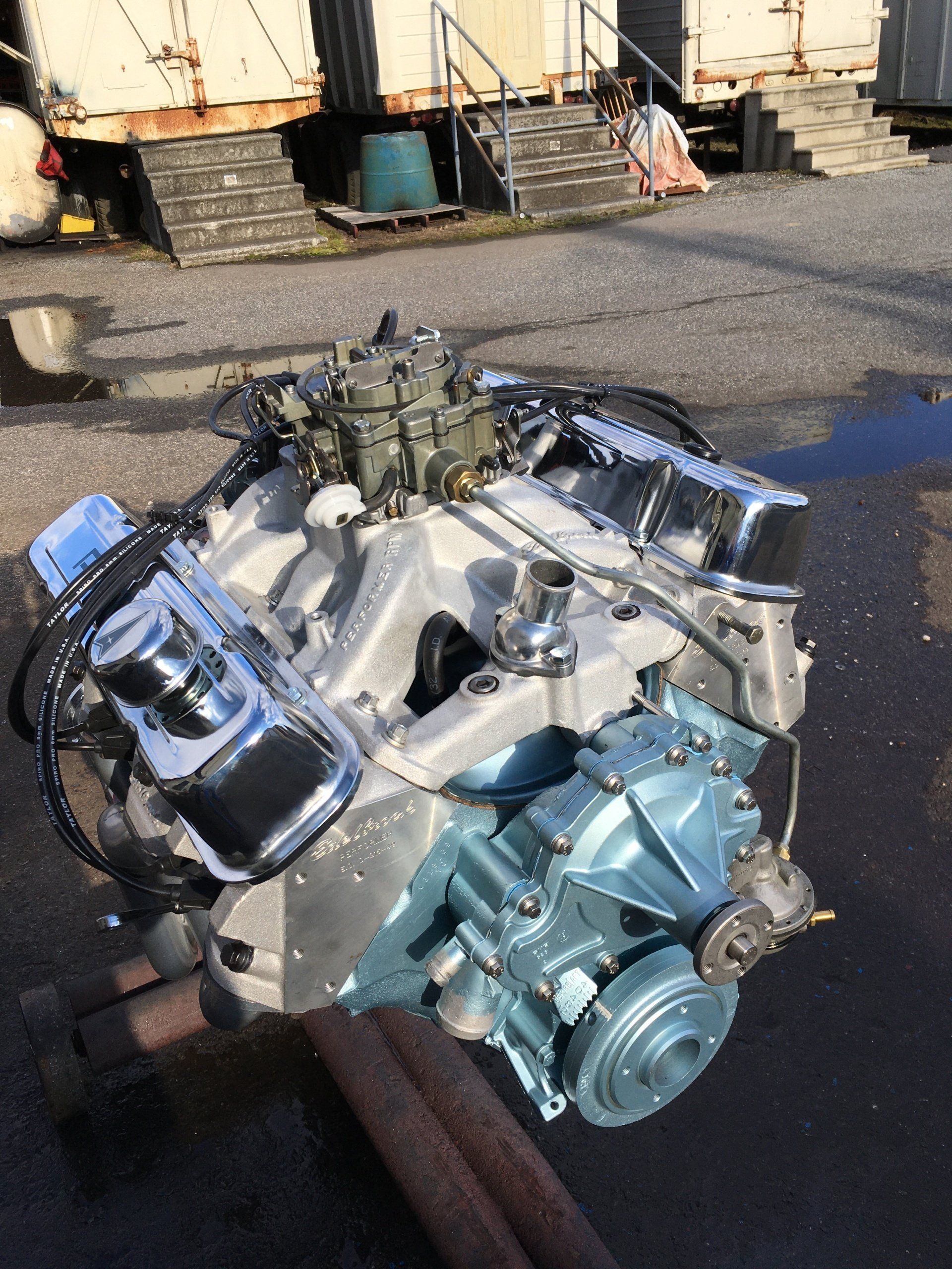 Pontiac engine, chrome valve covers and carburetor, blue painted transmission, on stand outside.