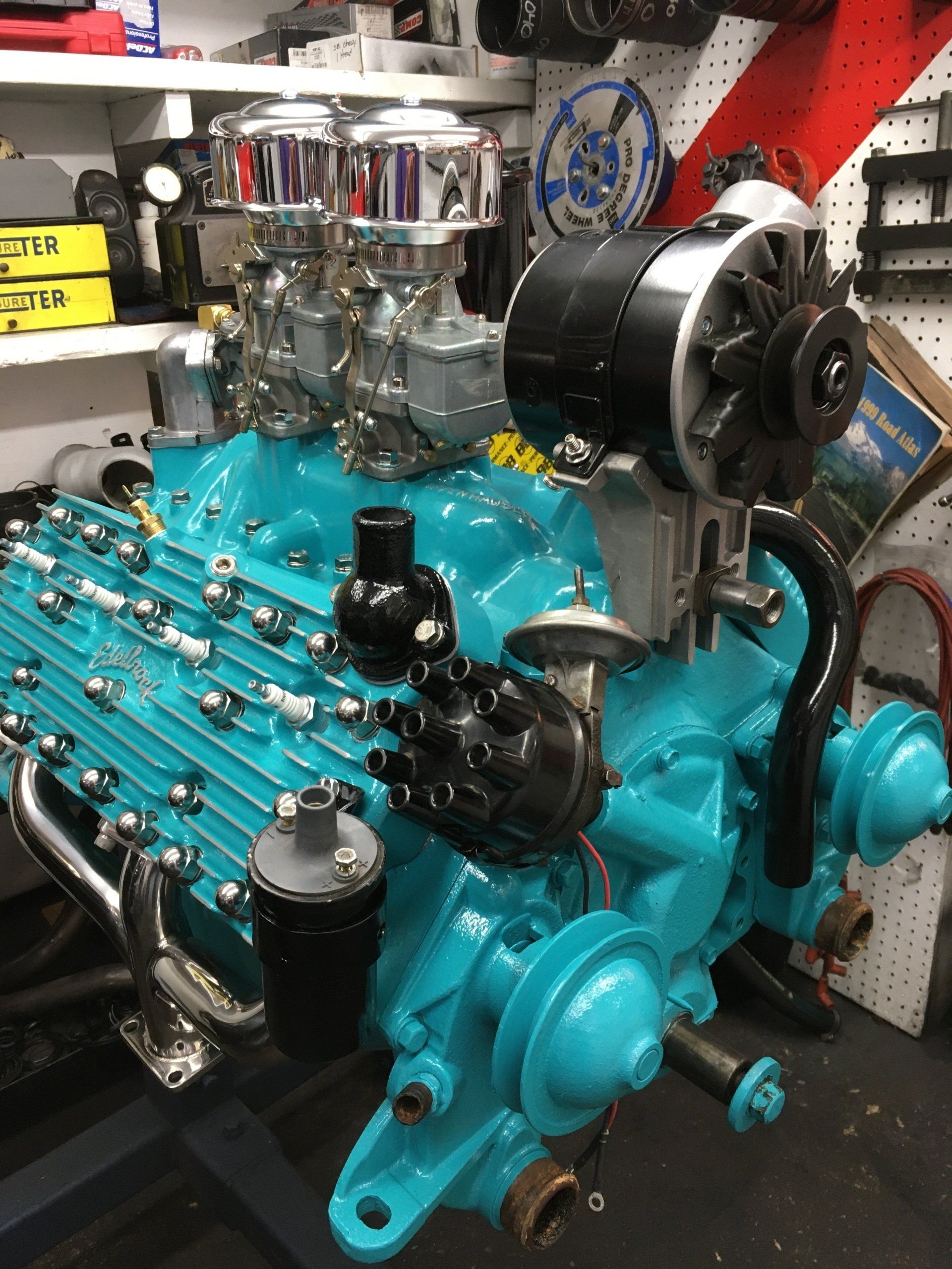 Turquoise car engine with chrome carburetors and black components in a workshop.