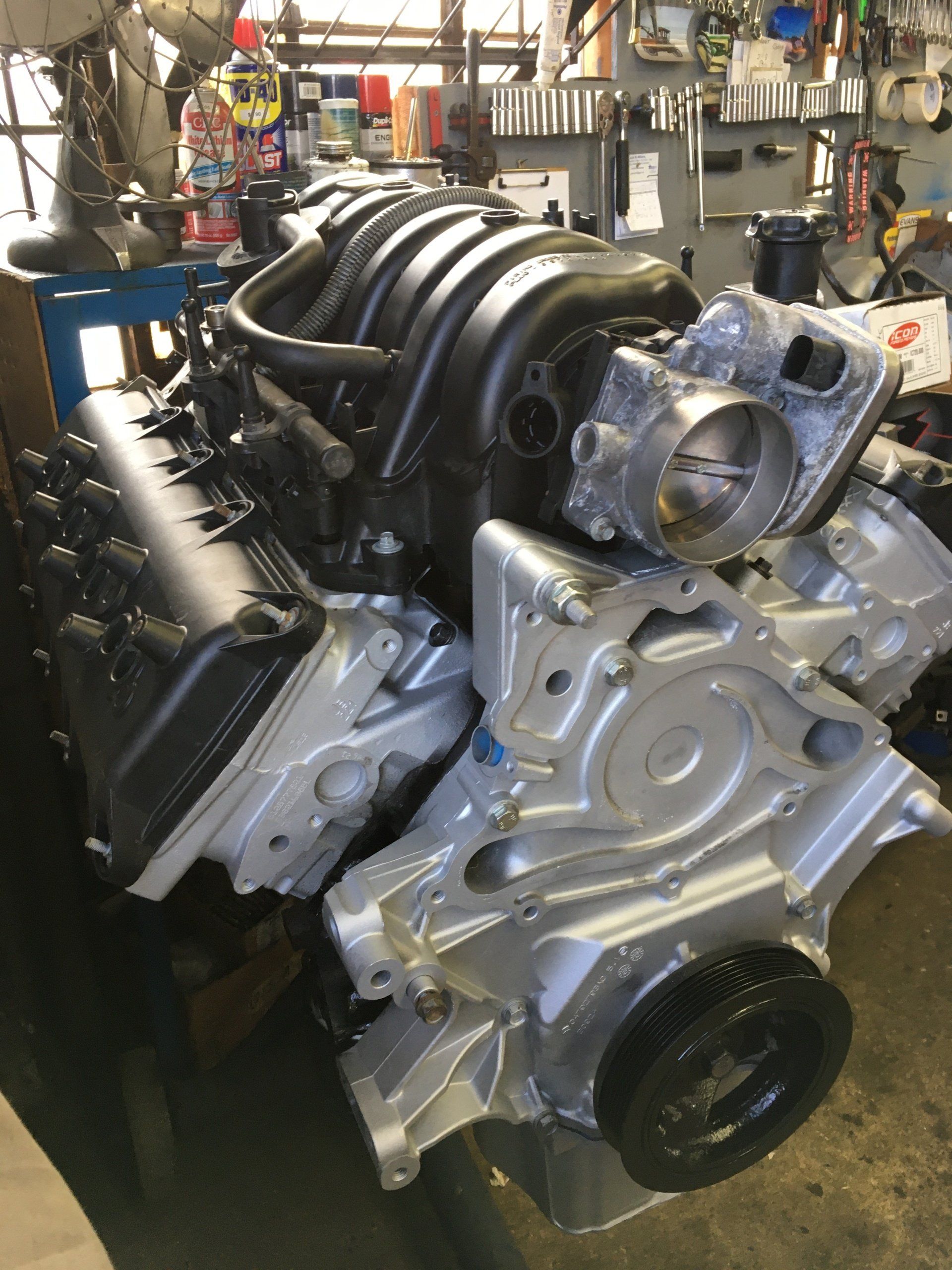Assembled car engine with black intake manifold, silver block and heads, sitting in a workshop.