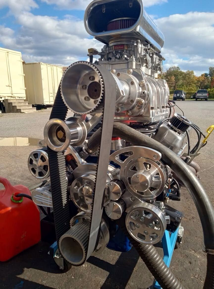 Supercharged engine with polished metal components and large belt, outside under a blue sky.