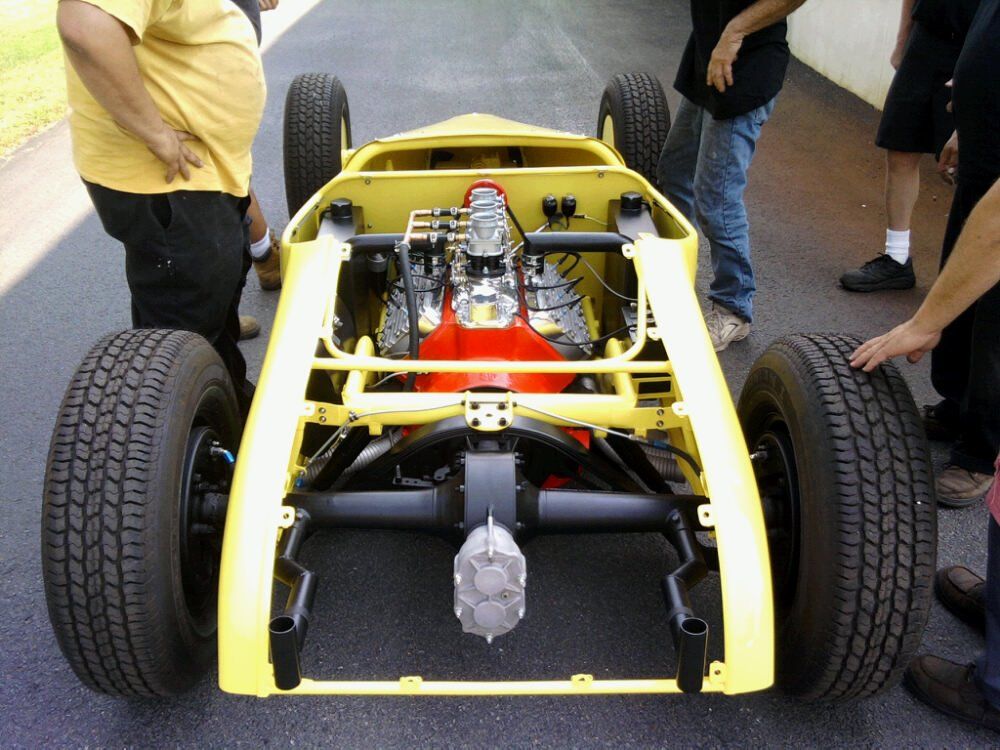 Yellow open-wheel hot rod chassis with engine, black wheels, and onlookers.